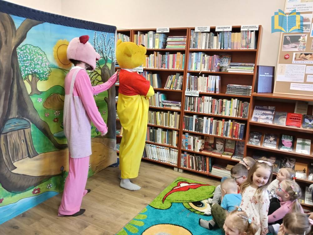 Zdjęcie: Spektakl teatralny w bibliotece. Kubuś Puchatek i Prosiaczek występują przed dziećmi. Za nimi, na płótnie kolorowy obraz wiosny.